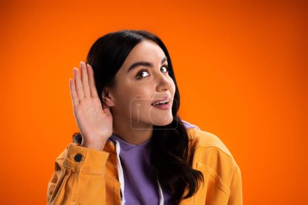 Téléchargez les photos : Young woman in jacket listening while holding hand near ear isolated on orange - en image libre de droit