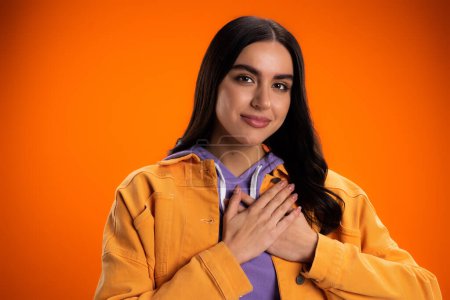 happy young woman touching chest isolated on orange