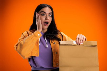 Shocked woman in jacket holding paper bag isolated on orange