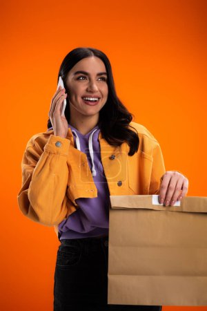 Cheerful brunette woman talking on smartphone and holding paper bag isolated on orange
