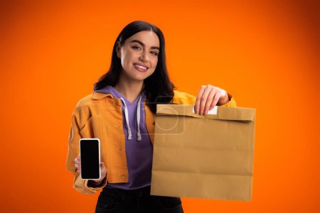Photo for Positive young woman showing smartphone and paper bag isolated on orange - Royalty Free Image