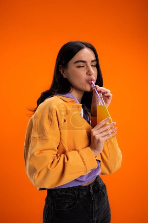 stylish woman holding glass bottle and drinking juice isolated on orange