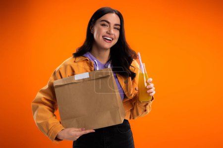 Cheerful young woman holding paper bag and juice in glass bottle isolated on orange