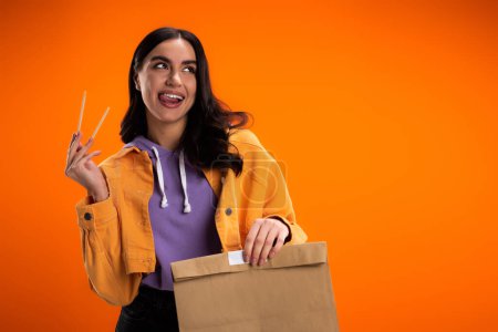 Joyful brunette woman holding chopsticks and paper bag isolated on orange