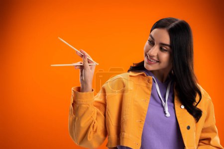 Young brunette woman looking at bamboo chopsticks isolated on orange