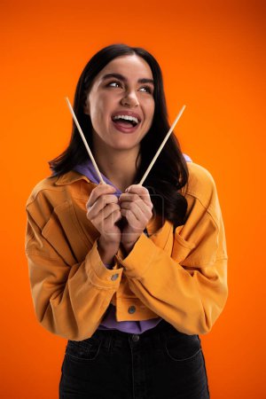Cheerful young woman holding bamboo chopsticks and looking away isolated on orange