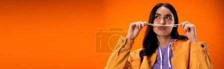 Pretty brunette woman holding chopsticks and pouting lips isolated on orange, banner 