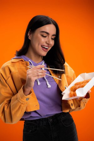 Positive brunette woman holding chopsticks and sushi in package isolated on orange