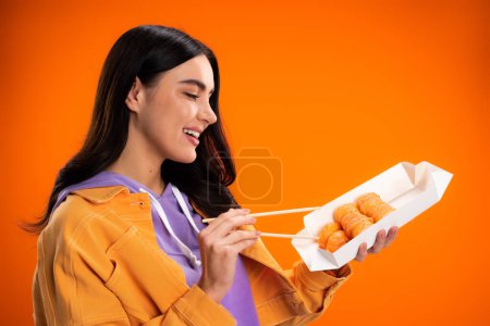 Smiling brunette woman holding takeaway sushi and chopsticks isolated on orange