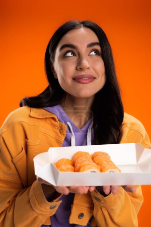 Portrait of dreamy young woman holding takeaway sushi isolated on orange