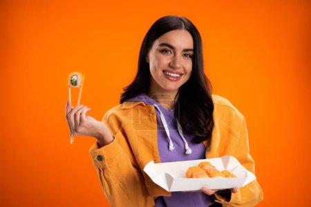Smiling brunette woman holding sushi and looking at camera isolated on orange