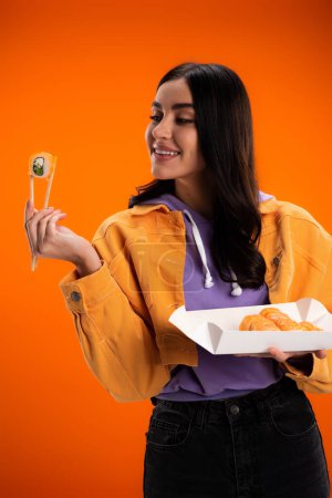 Stylish young woman looking at sushi and chopsticks isolated on orange