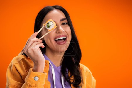 Positive young woman holding sushi in chopsticks near eye isolated on orange
