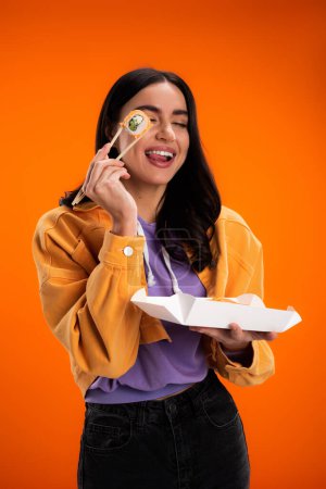 Positive woman holding takeaway sushi and chopsticks while sticking out tongue isolated on orange