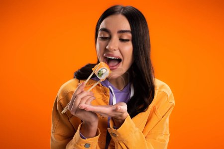 Brunette woman opening mouth and holding sushi isolated on orange