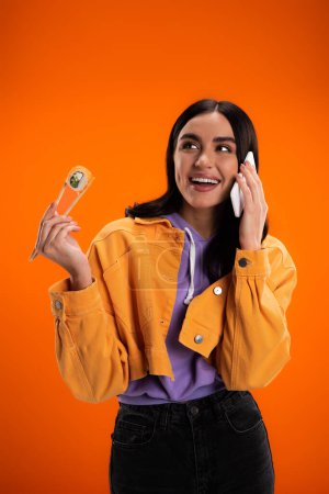 Positive brunette woman holding sushi in chopsticks and talking on smartphone isolated on orange