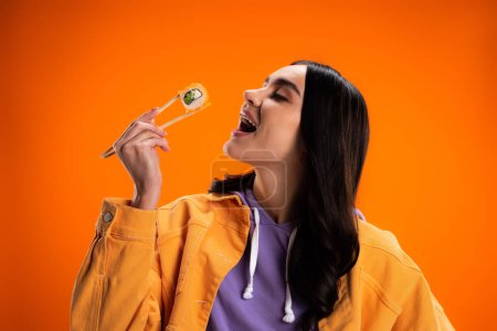 Stylish brunette woman opening mouth and holding sushi isolated on orange