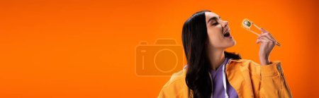 Young woman opening mouth and holding sushi in chopsticks isolated on orange, banner 