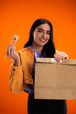 happy brunette woman with paper bag and sushi roll looking at camera isolated on orange