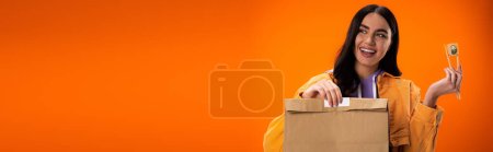 positive brunette woman with paper bag and fresh sushi roll looking away isolated on orange, banner