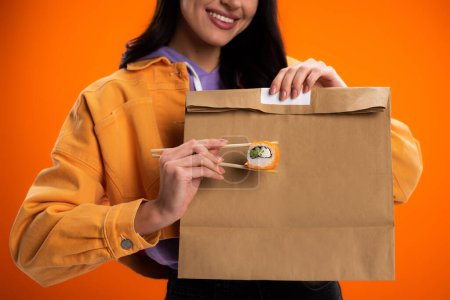cropped view of smiling woman with sushi roll and paper bag isolated on orange