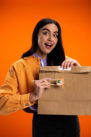 amazed woman holding chopsticks with sushi roll near paper bag isolated on orange