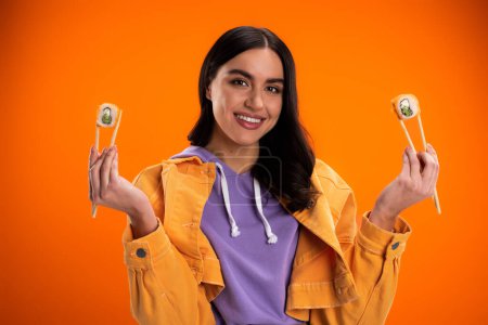 Téléchargez les photos : Young brunette woman smiling at camera while holding chopsticks with sushi rolls isolated on orange - en image libre de droit