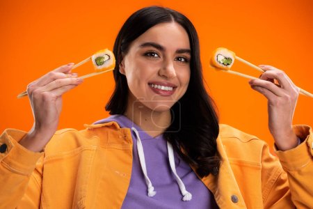 happy brunette woman in jacket and purple hoodie posing with delicious sushi rolls isolated on orange