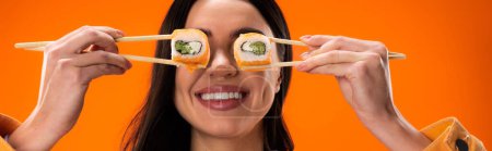 cheerful brunette woman covering eyes with fresh sushi rolls isolated on orange, banner