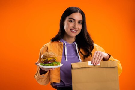 Téléchargez les photos : Happy brunette woman holding paper bag and delicious burger isolated on orange - en image libre de droit