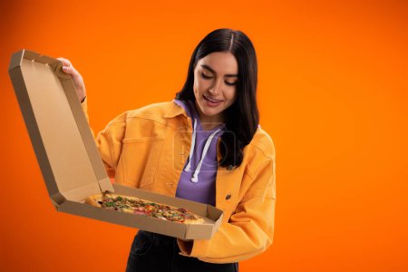 Foto de Pleased woman in trendy jacket looking at tasty pizza in cardboard box isolated on orange - Imagen libre de derechos