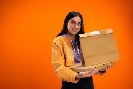 brunette woman with pizza box and paper bag smiling at camera isolated on orange