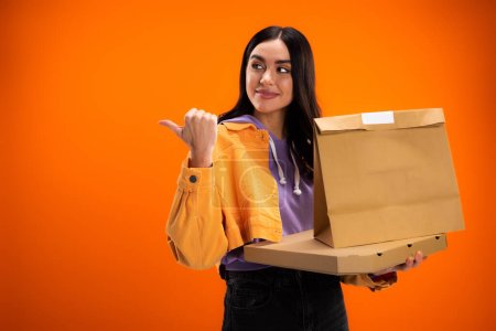 young woman with paper bag and pizza box looking away and pointing with thumb isolated on orange