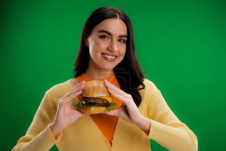 happy and stylish woman holding tasty burger and looking at camera isolated on green