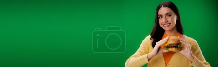 smiling woman with tasty burger looking at camera isolated on green, banner