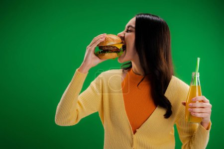 brunette woman holding bottle of fresh lemonade and eating tasty burger isolated on green