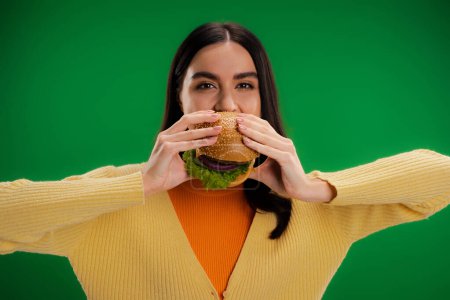 young brunette woman eating delicious burger and looking at camera isolated on green