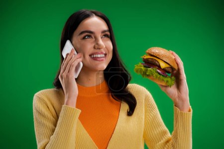 happy young woman holding delicious burger and talking on smartphone isolated on green
