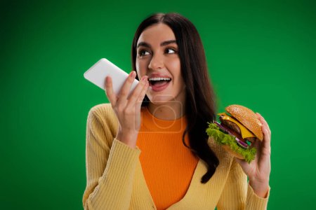 happy woman holding tasty burger and sending voice message on smartphone isolated on green