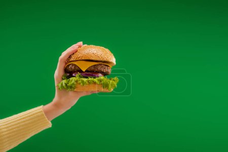 Foto de Partial view of woman holding tasty burger with lettuce and cheese with meat isolated on green - Imagen libre de derechos