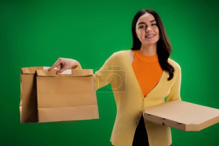 smiling woman holding craft paper bags and pizza box while looking at camera isolated on green