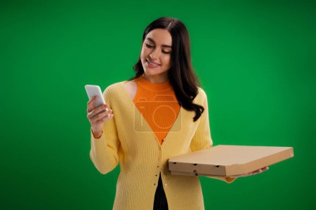 brunette woman holding cardboard pizza box and using mobile phone isolated on green