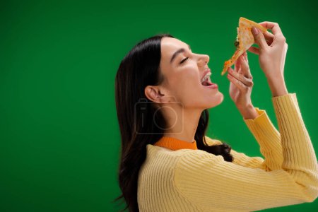 Téléchargez les photos : Side view of young and hungry woman eating piece of pizza isolated on green - en image libre de droit