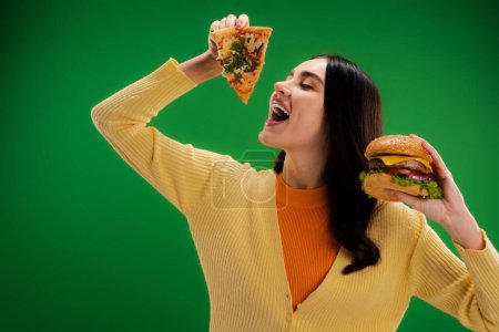 hungry woman holding delicious burger and opening mouth near piece of pizza isolated on green