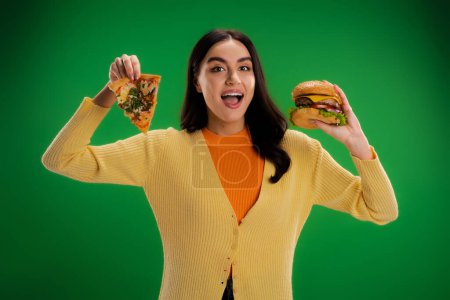 young amazed woman with piece of pizza and tasty burger looking at camera isolated on green