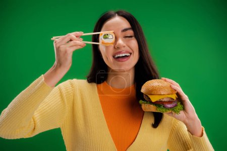cheerful woman with tasty burger holding sushi roll near eye isolated on green