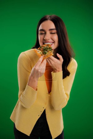 pleased woman with closed eyes eating delicious pizza isolated on green