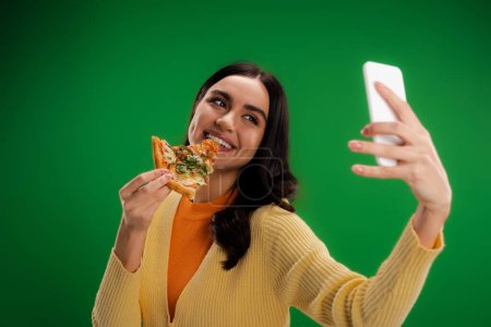 cheerful woman biting piece of pizza and taking selfie on smartphone isolated on green