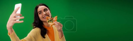 smiling brunette woman taking selfie on cellphone while eating tasty pizza isolated on green, banner