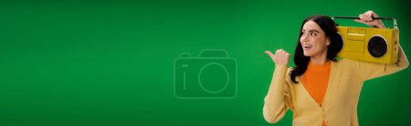 Foto de Cheerful brunette woman holding boombox and pointing with thumb isolated on green, banner - Imagen libre de derechos